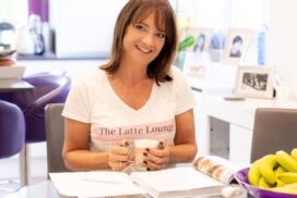 Katie sits at a table with a latte and a book, wearing a Latte Lounge t-shirt and smiling at the camera