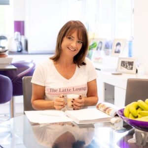 Katie sits at a table with a latte and a book, wearing a Latte Lounge t-shirt and smiling at the camera